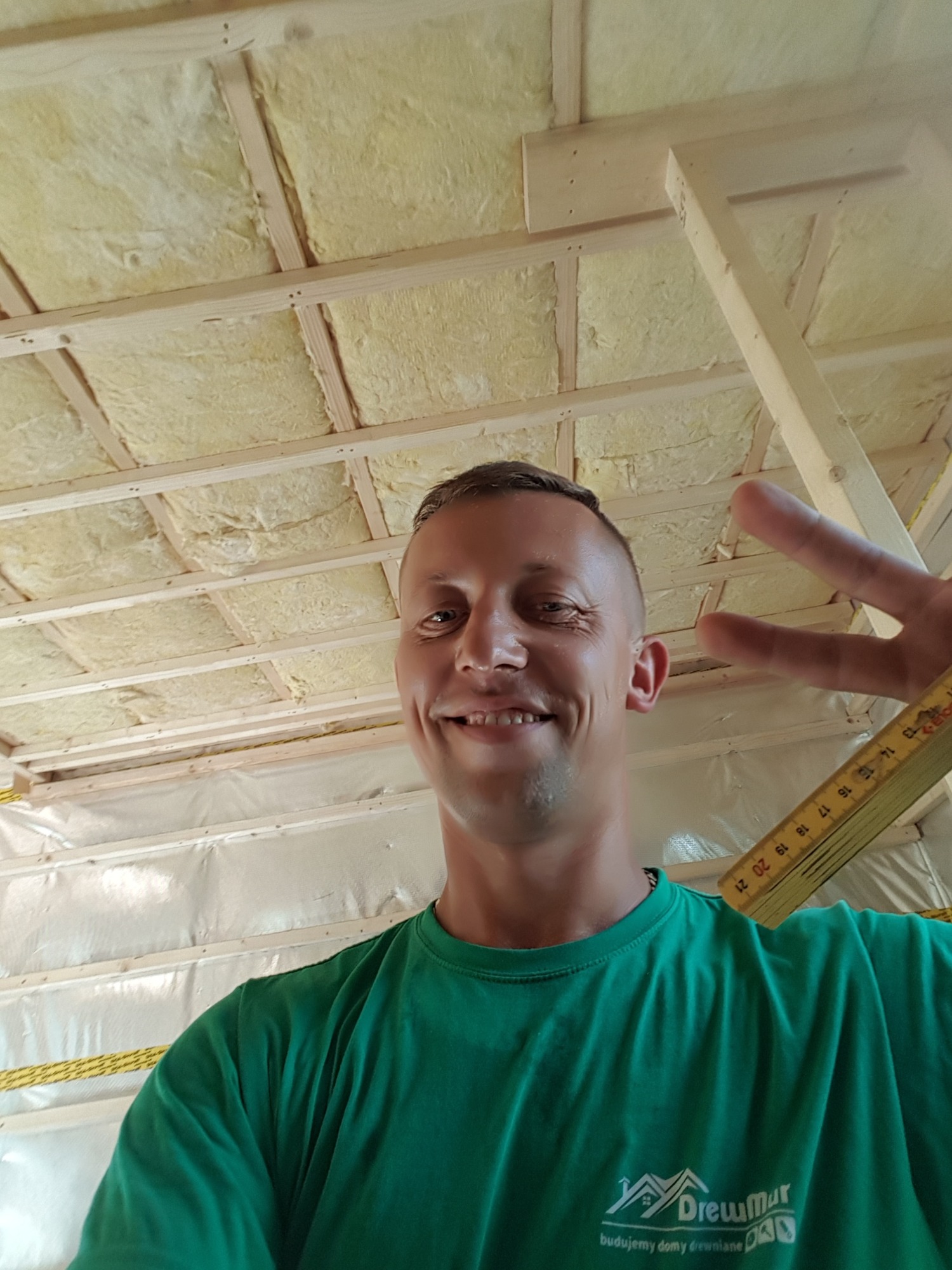 Uśmiechnięty mężczyzna w zielonej koszulce z logo firmy budowlanej, robiący selfie na tle konstrukcji dachu z izolacją z wełny mineralnej i folią paroizolacyjną, trzymający rozłożoną miarkę stolarską.