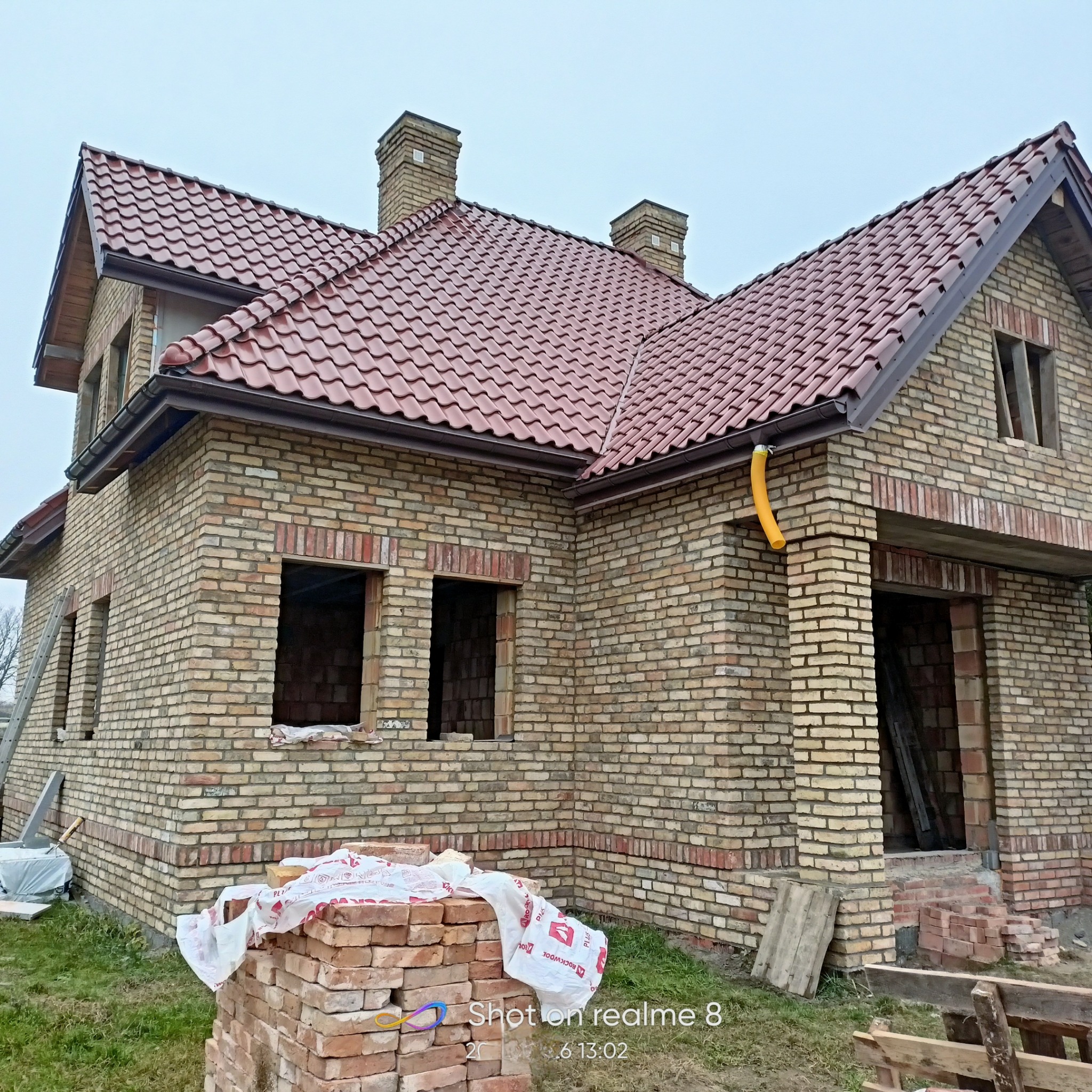 Wznoszony budynek z cegły klinkierowej z czerwoną dachówką, widoczne niezamurowane otwory okienne i drzwiowe, stos cegieł na pierwszym planie, żółta rura spustowa przy elewacji.