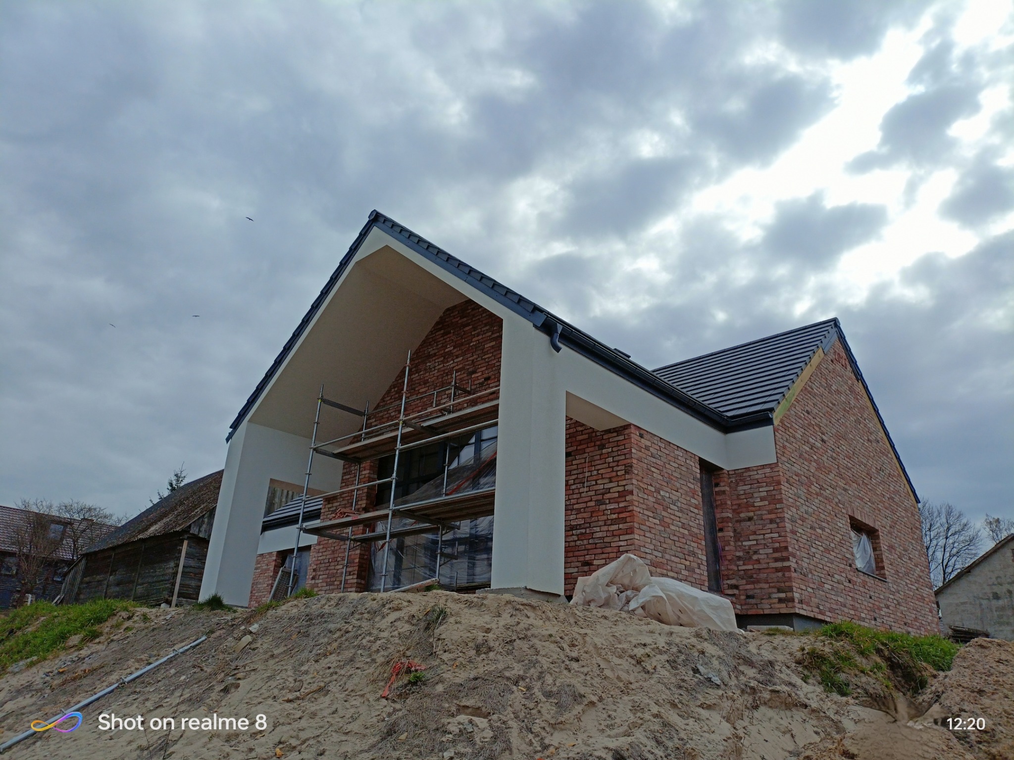 Dom jednorodzinny w trakcie budowy elewacji z cegły klinkierowej, widoczne rusztowania i zabezpieczone okna, pochmurne niebo w tle.