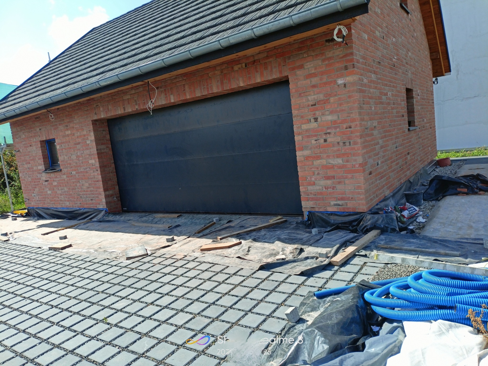 Surowa bryła garażu z czerwonej cegły z czarną bramą segmentową, widoczne elementy instalacji elektrycznej, otoczenie w trakcie prac wykończeniowych, ułożona kostka ażurowa.