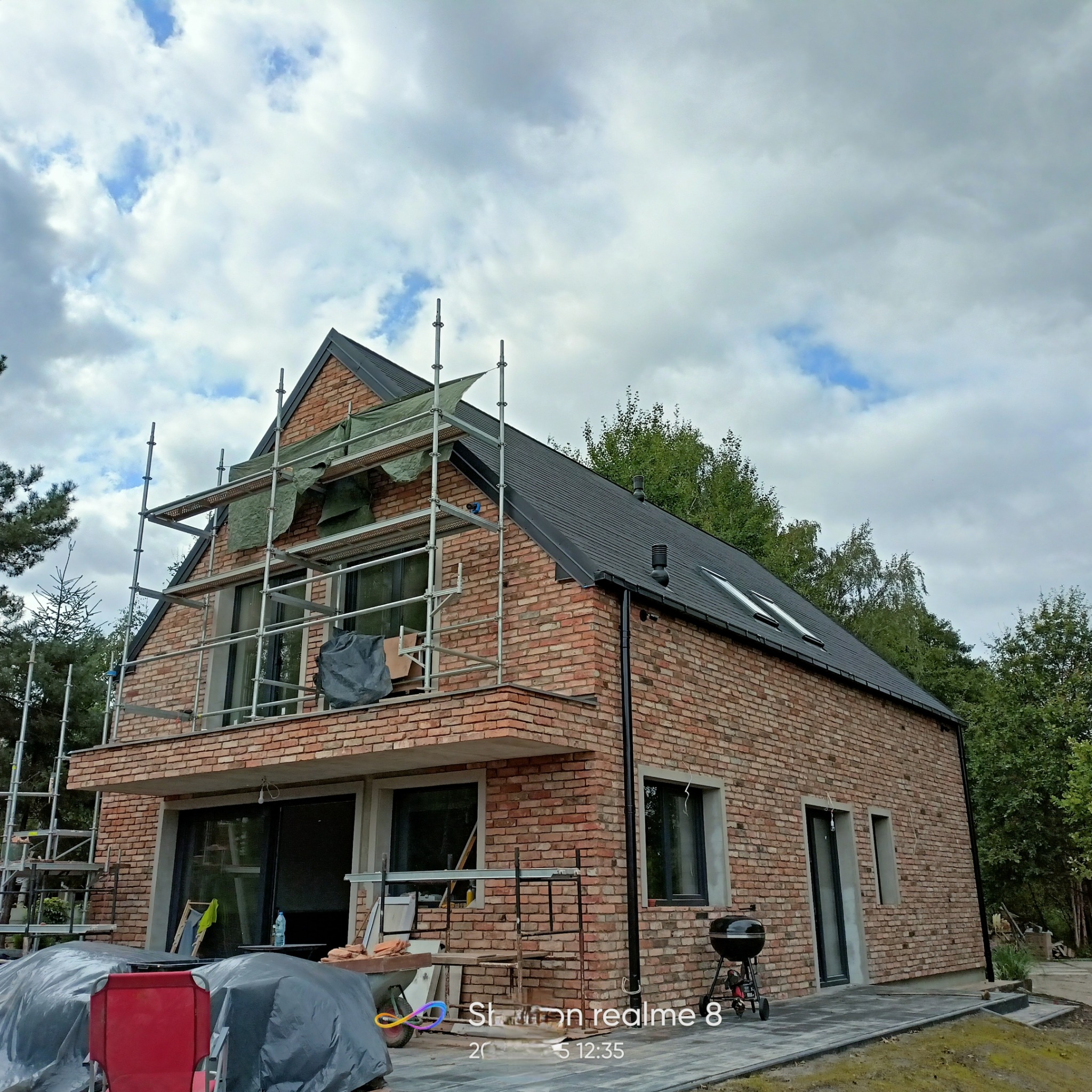 Dwukondygnacyjny dom z cegły z czarnym dachem, częściowo otoczony rusztowaniem, z widocznymi narzędziami i materiałami budowlanymi na zewnątrz.