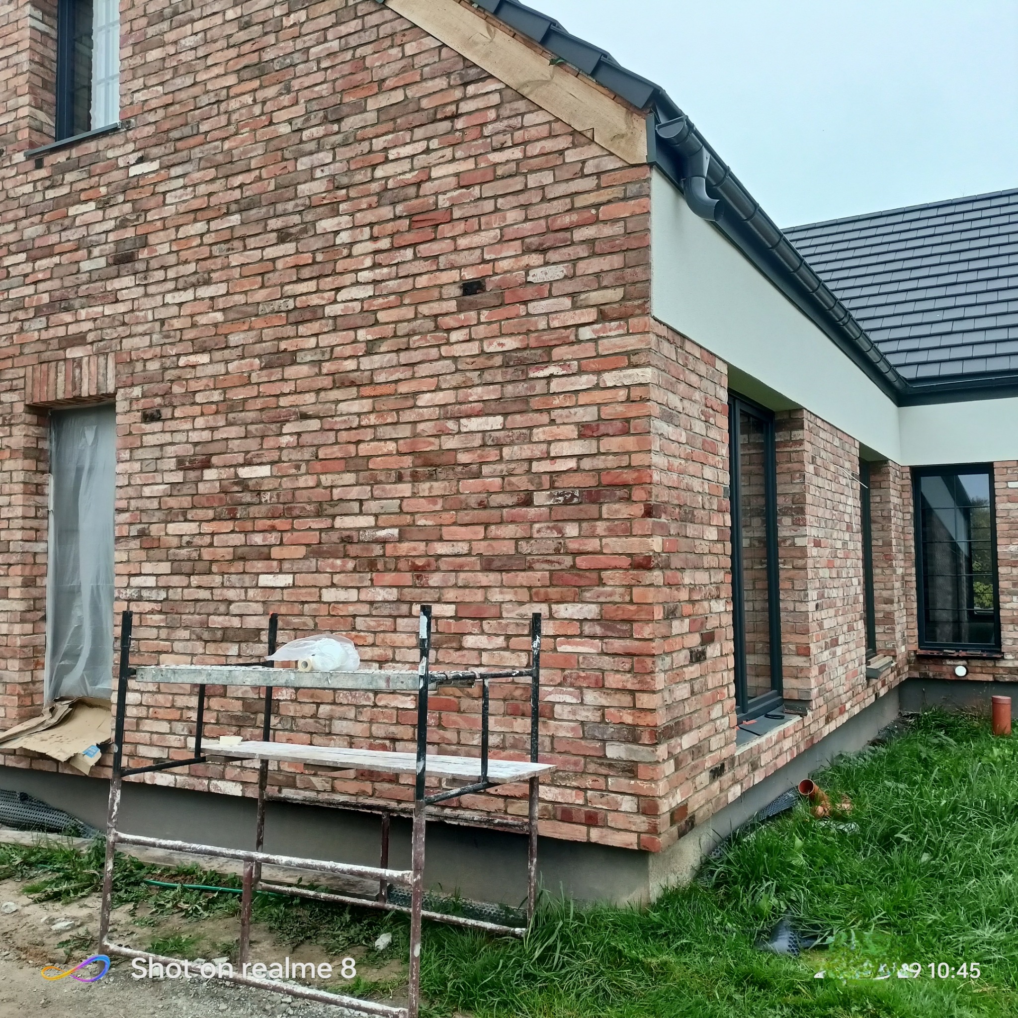 Murowana elewacja budynku z cegły klinkierowej w trakcie budowy, widoczne rusztowanie, okna z czarnymi ramami i fragment dachu z grafitową dachówką.
