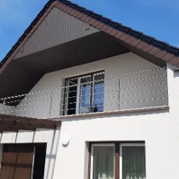 Biały dom z balkonem zabezpieczonym minimalistyczną balustradą nierdzewną w Zbiersku-Cukrowni, widok na błękitne niebo z lekkimi obłokami.