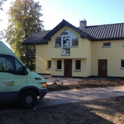 Żółty dom murowany z dwuspadowym dachem z ciemnej dachówki, widoczny z boku wraz z fragmentem firmowego samochodu z logo budowlanym na tle drzew i brukowanego podjazdu.