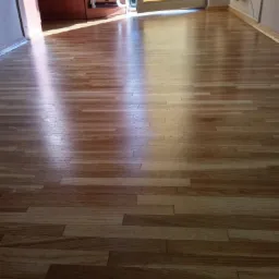 Wycyklinowana i polakierowana podłoga parkietowa z wyraźnym usłojeniem, odbijająca światło słoneczne wpadające przez okno z żaluzjami.