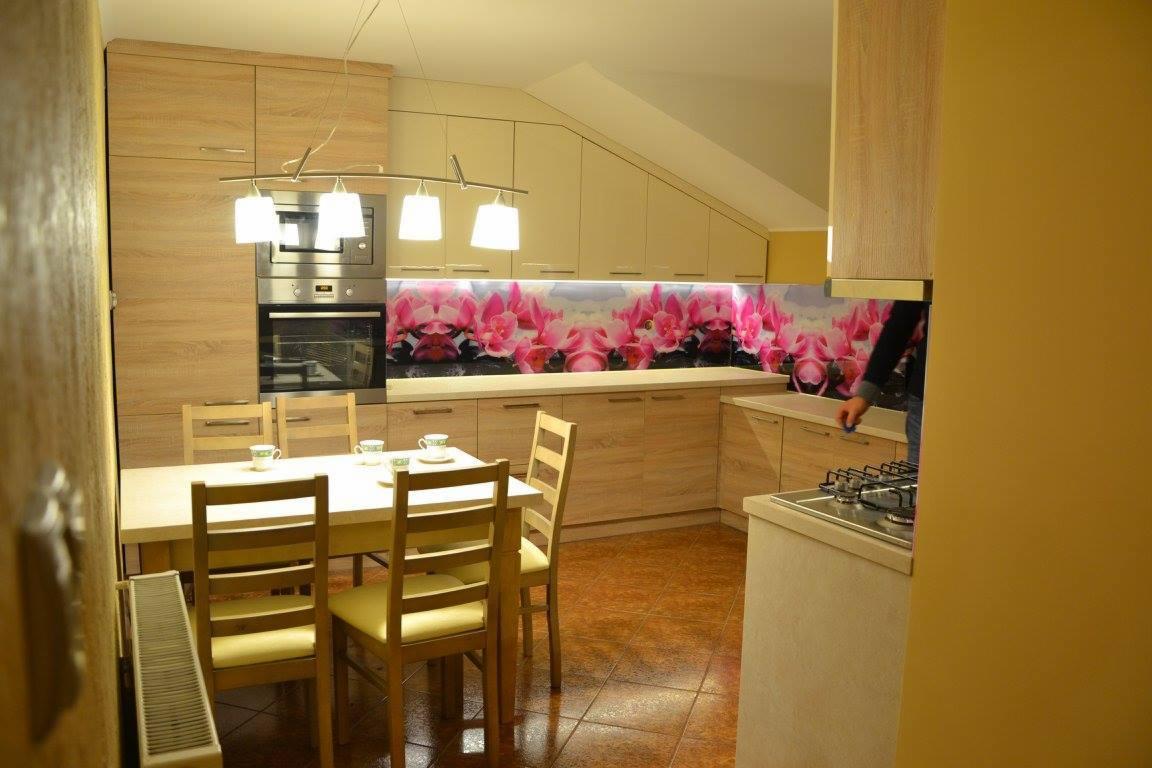 Jasna kuchnia z drewnianymi meblami i szklanym panelem ściennym z motywem kwiatowym, widoczne krzesła przy stole z filiżankami.