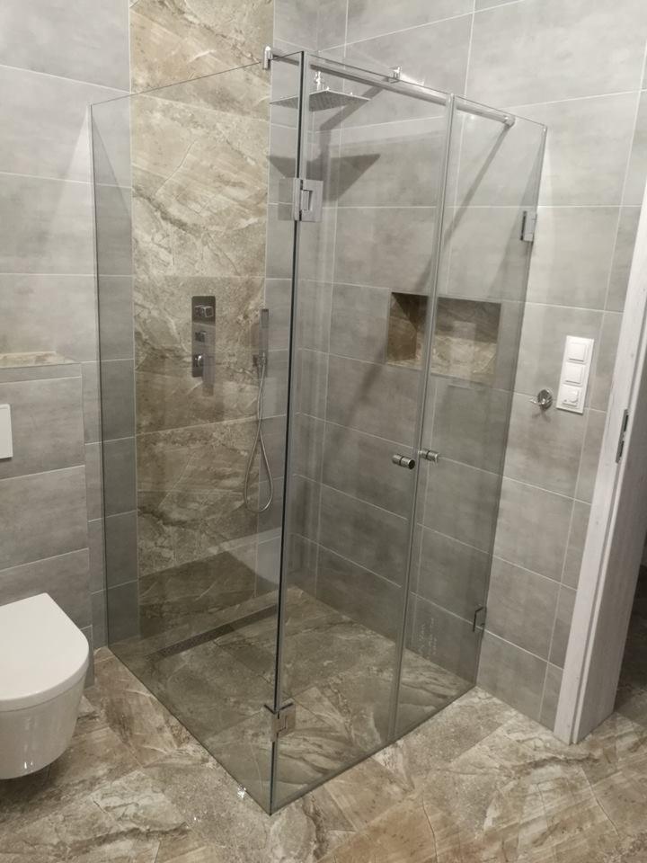 Kabina prysznicowa z przezroczystego szkła, zamontowana w rogu łazienki wyłożonej szarymi i beżowymi płytkami imitującymi marmur, widoczny biały sedes po lewej stronie.