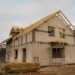 Surowy stan domu w trakcie budowy z pustak&oacute;w, z drewnianą konstrukcją dachu, z widocznymi rusztowaniami i stosem desek na pierwszym planie.