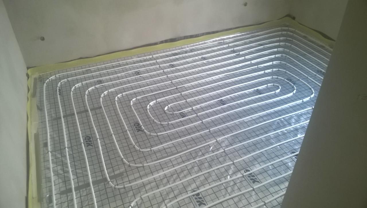 Białe rury ogrzewania podłogowego ułożone w pętle na metalowej siatce, przyklejone taśmą do ściany, przygotowane do zalania wylewką.