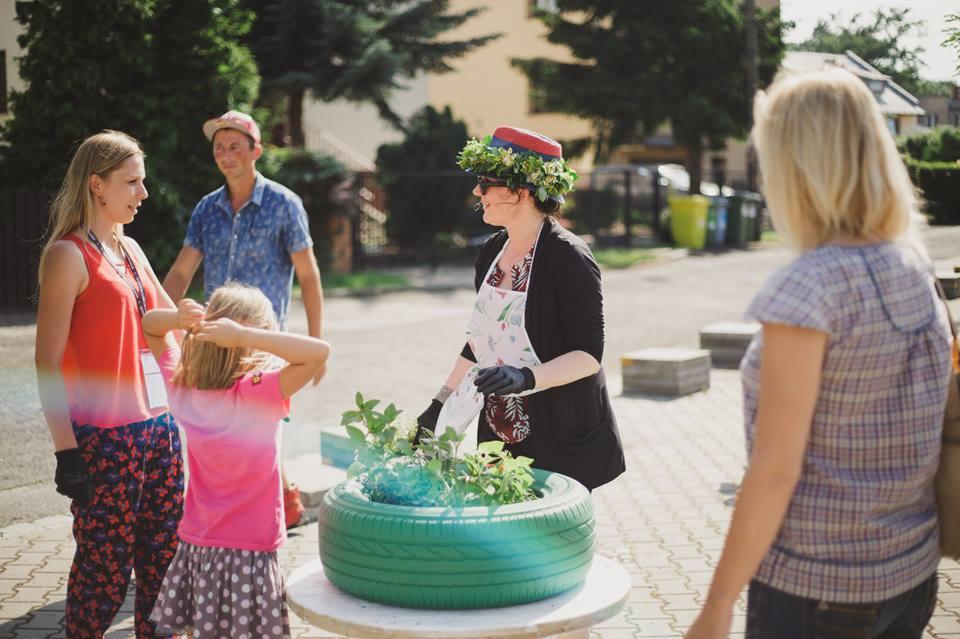 Kobieta w wianku i fartuchu sadzi rośliny w pomalowanej na zielono oponie samochodowej, wokół stoją inne osoby.
