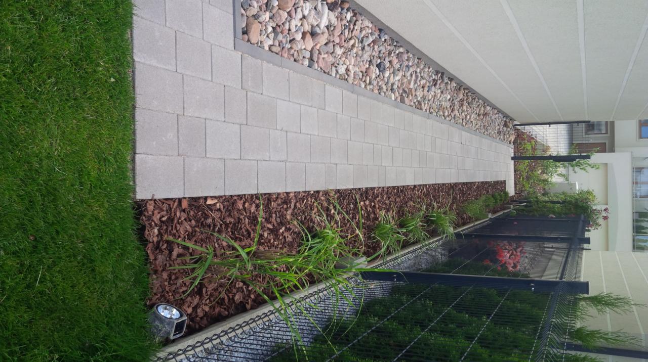 Ścieżka z szarych kostek brukowych, obok trawnik z lampą najazdową, rabata z kory i traw oraz pas z kamieni ozdobnych wzdłuż budynku.