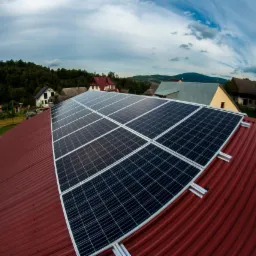 Panele na dachu z blachy trapezowej w odcieniach czerwieni, widok z góry na okoliczne domy i zalesione wzgórza, pochmurne niebo.