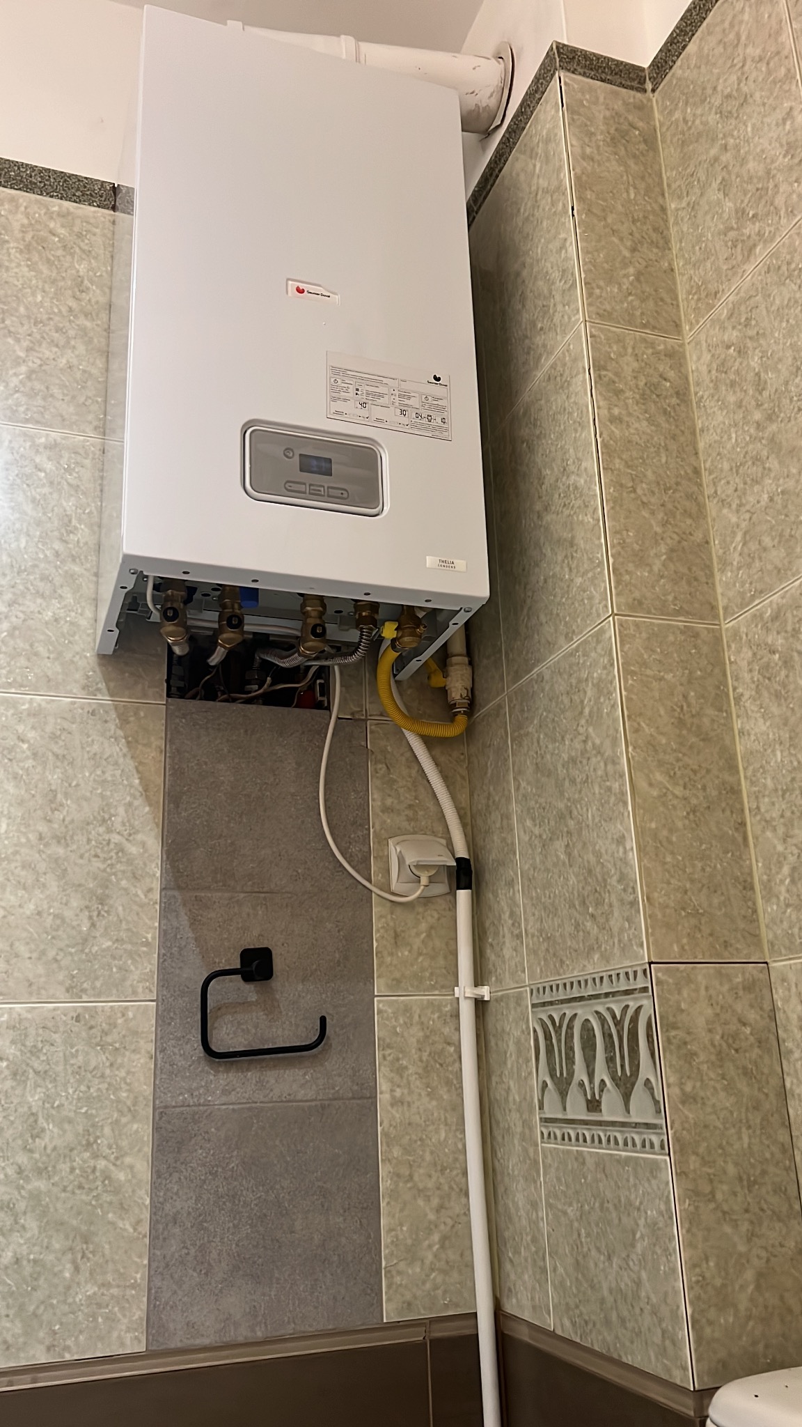 Biały piec gazowy zamontowany na ścianie z beżowych płytek. Widoczne przyłącza gazowe i hydrauliczne. U dołu uchwyt na papier toaletowy.