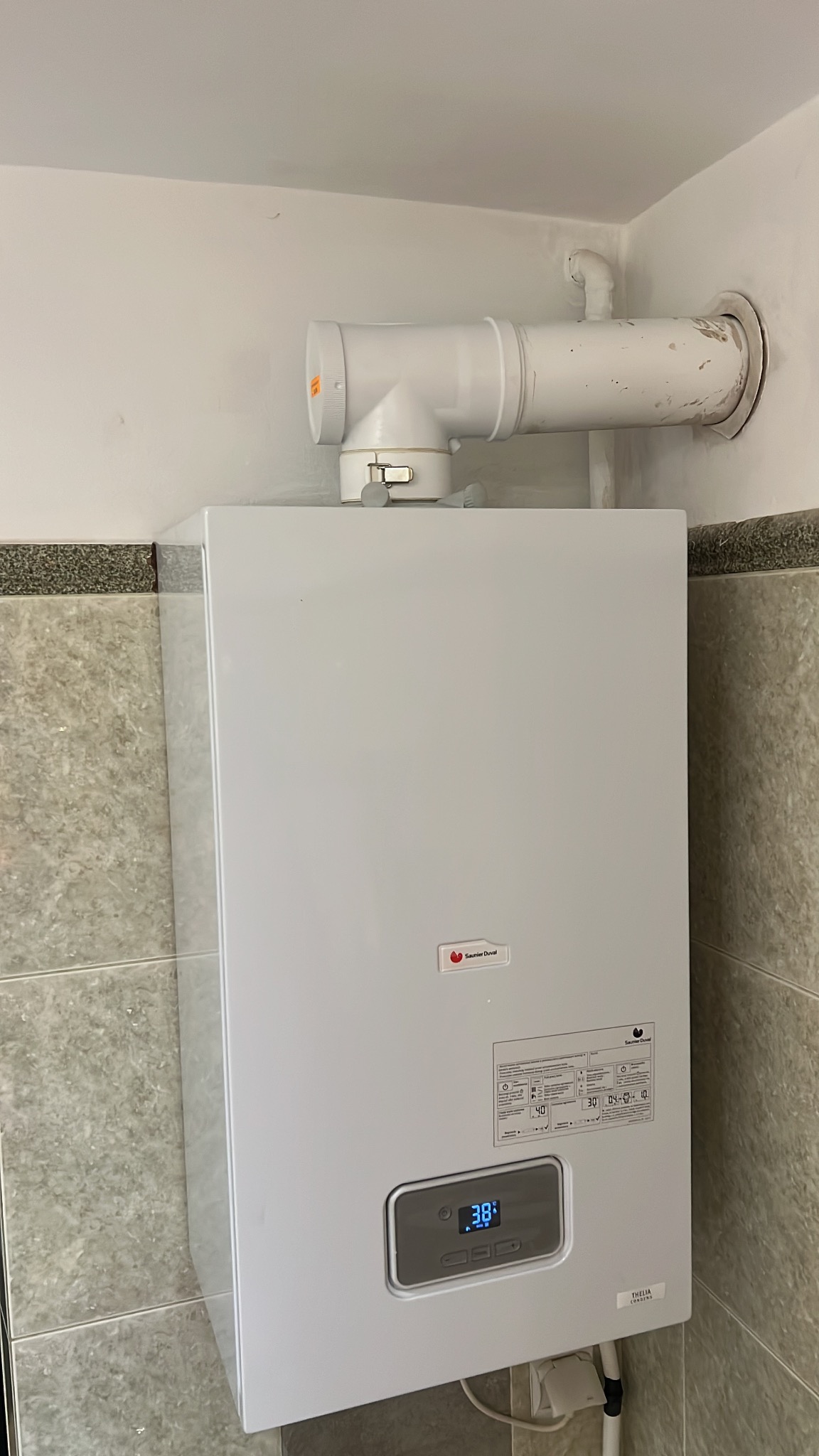 Biały, wiszący piec gazowy Saunier Duval z widocznym wyświetlaczem temperatury 38 stopni Celsjusza i rurą odprowadzającą spaliny, zamontowany na tle płytek.