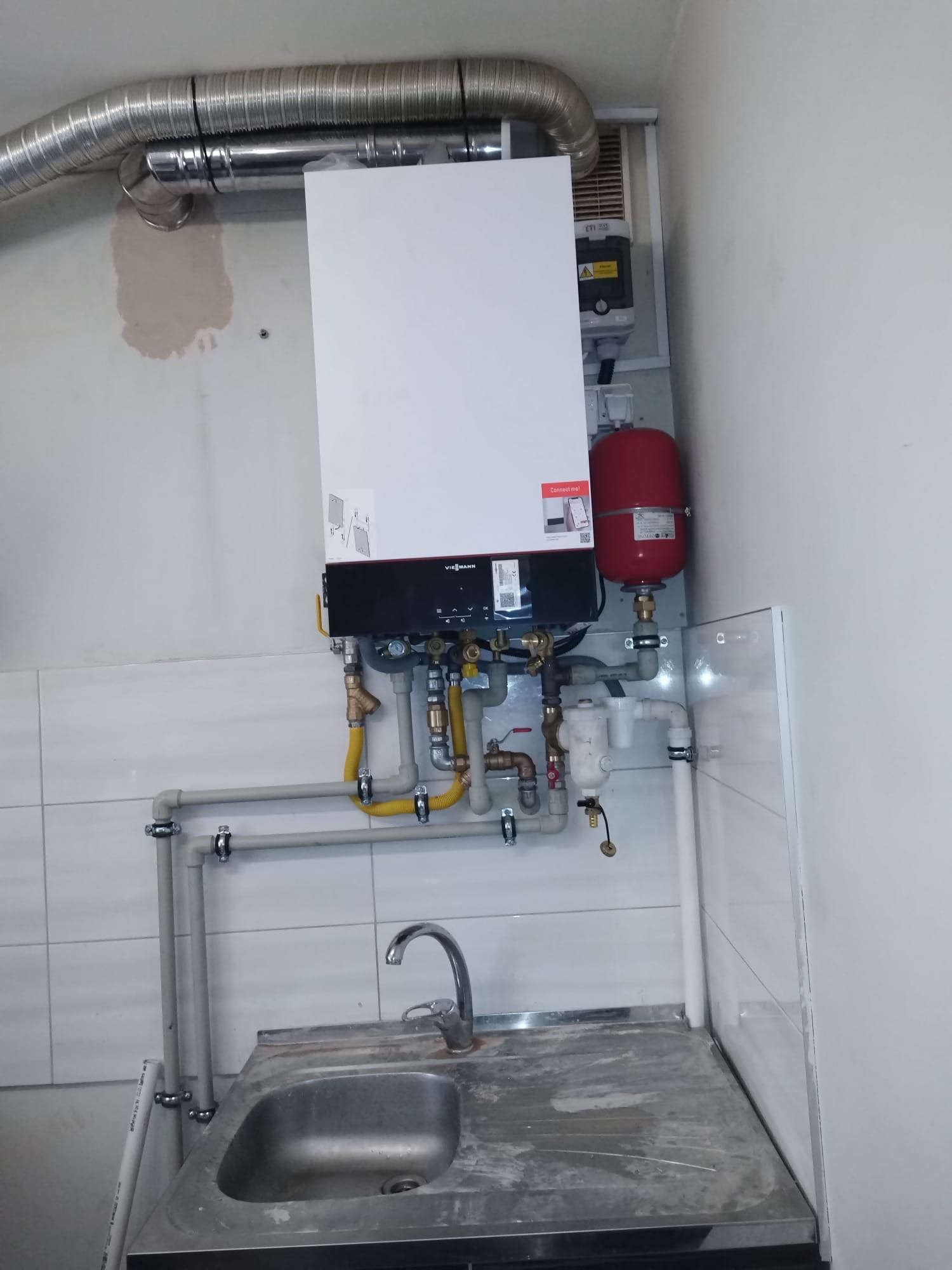 Zamontowany piec gazowy Viessmann z podłączeniami hydraulicznymi i odprowadzeniem spalin, widoczny nad zlewem w pomieszczeniu wykończonym białymi płytkami.