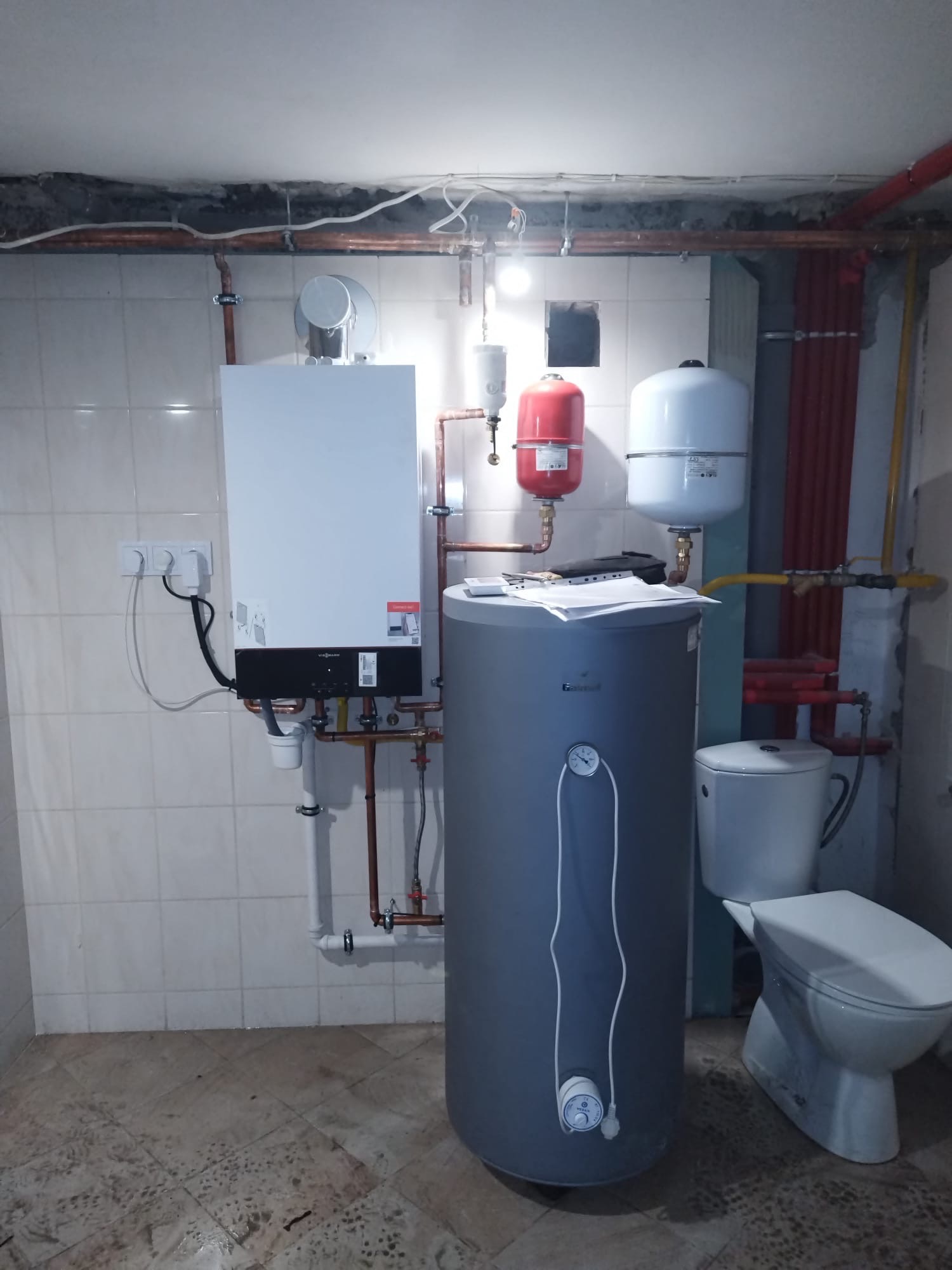 Biały piec gazowy Viessmann, szary zasobnik wody, czerwony i biały zbiornik wyrównawczy, rury miedziane na tle białych płytek. Widoczna toaleta.