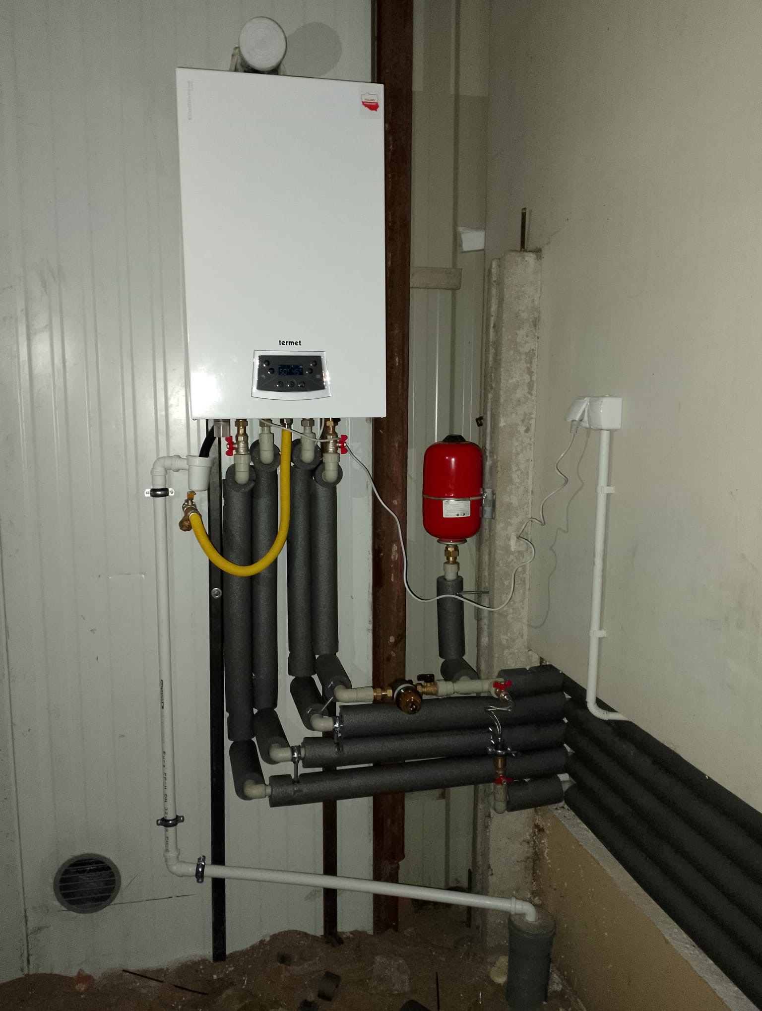 Biały piec gazowy Termet zamontowany na ścianie, z widocznymi rurami w izolacji, żółtym przewodem gazowym i czerwonym zbiornikiem wyrównawczym. Instalacja w surowym pomieszczeniu.