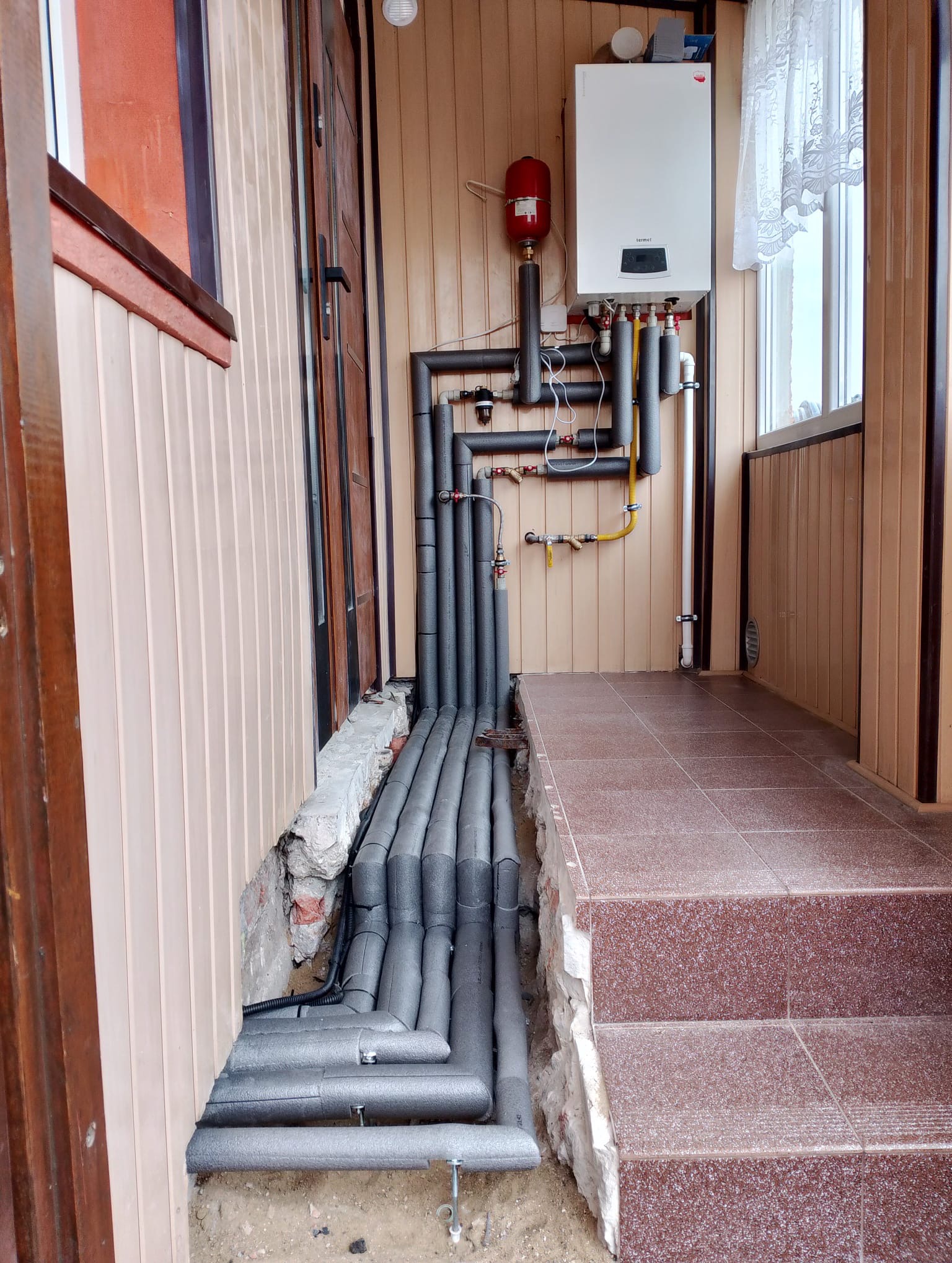 Instalacja grzewcza z piecem gazowym na zewnątrz budynku, rury zaizolowane szarą pianką, widoczny zbiornik wyrównawczy i fragment okna z firanką.