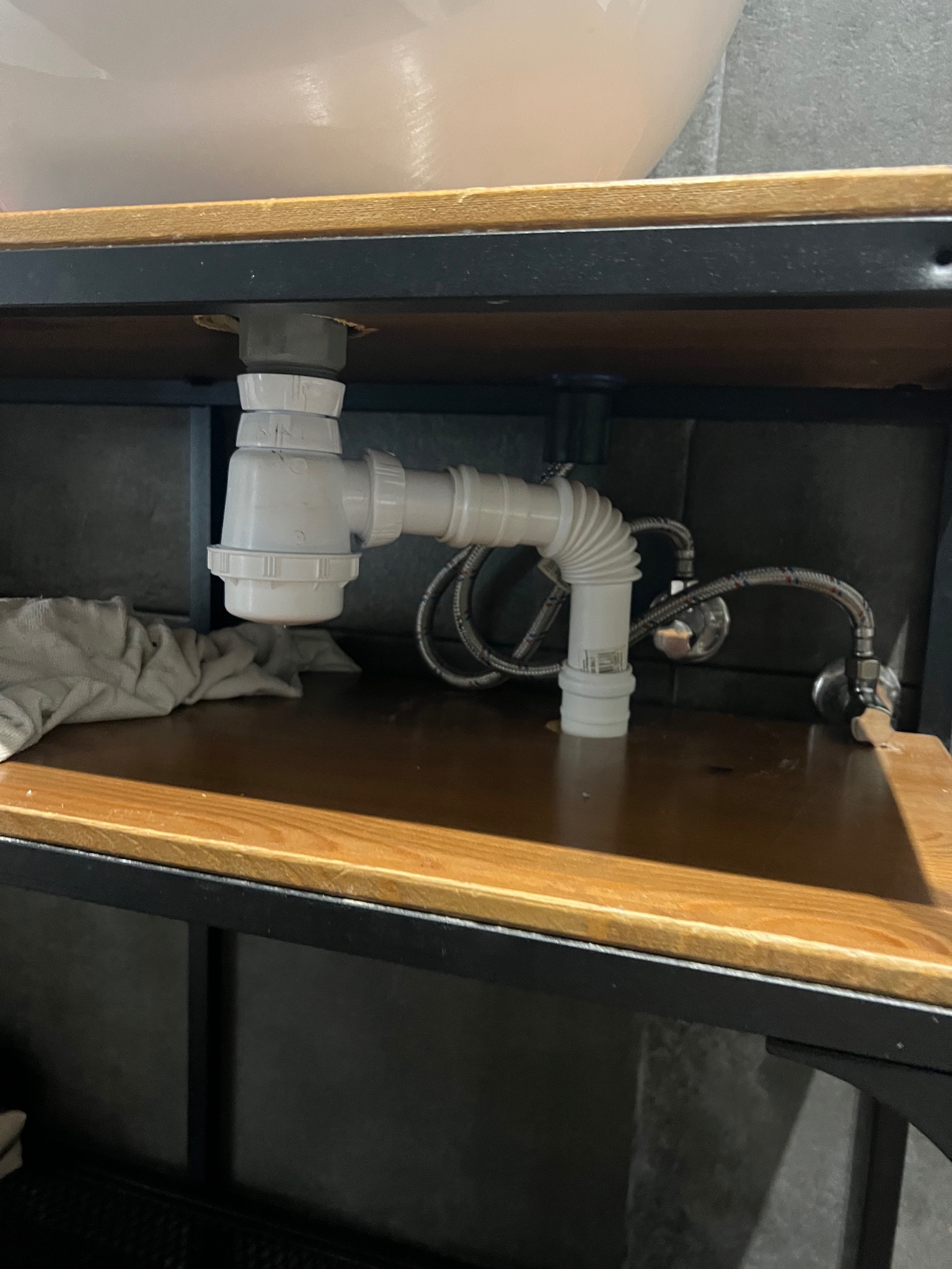 Biały syfon umywalkowy z elastycznym wężem pod drewnianą szafką, widoczne zawory i szara ściana w tle. Ujęcie z dołu, podkreślające instalację hydrauliczną.