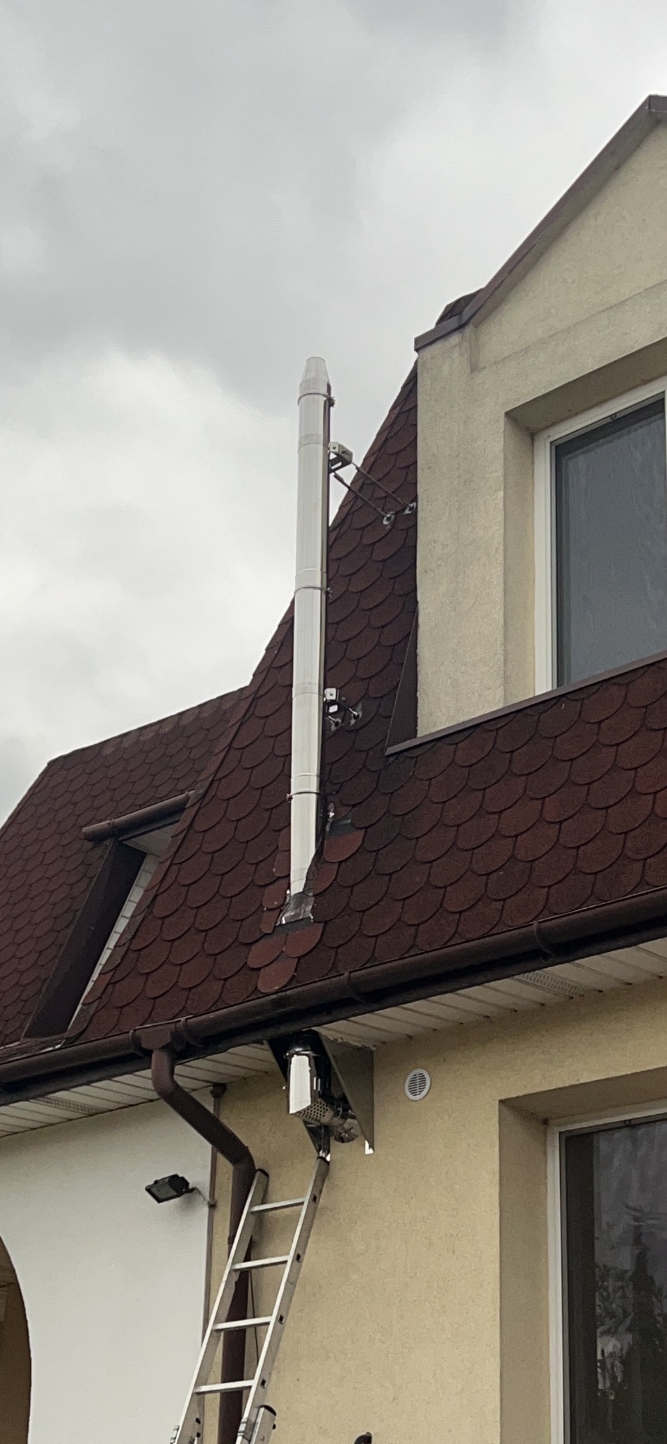 Montaż komina systemowego ze stali nierdzewnej na dachu z dachówki bitumicznej. Widoczna drabina oparta o ścianę budynku. Pochmurne niebo w tle.