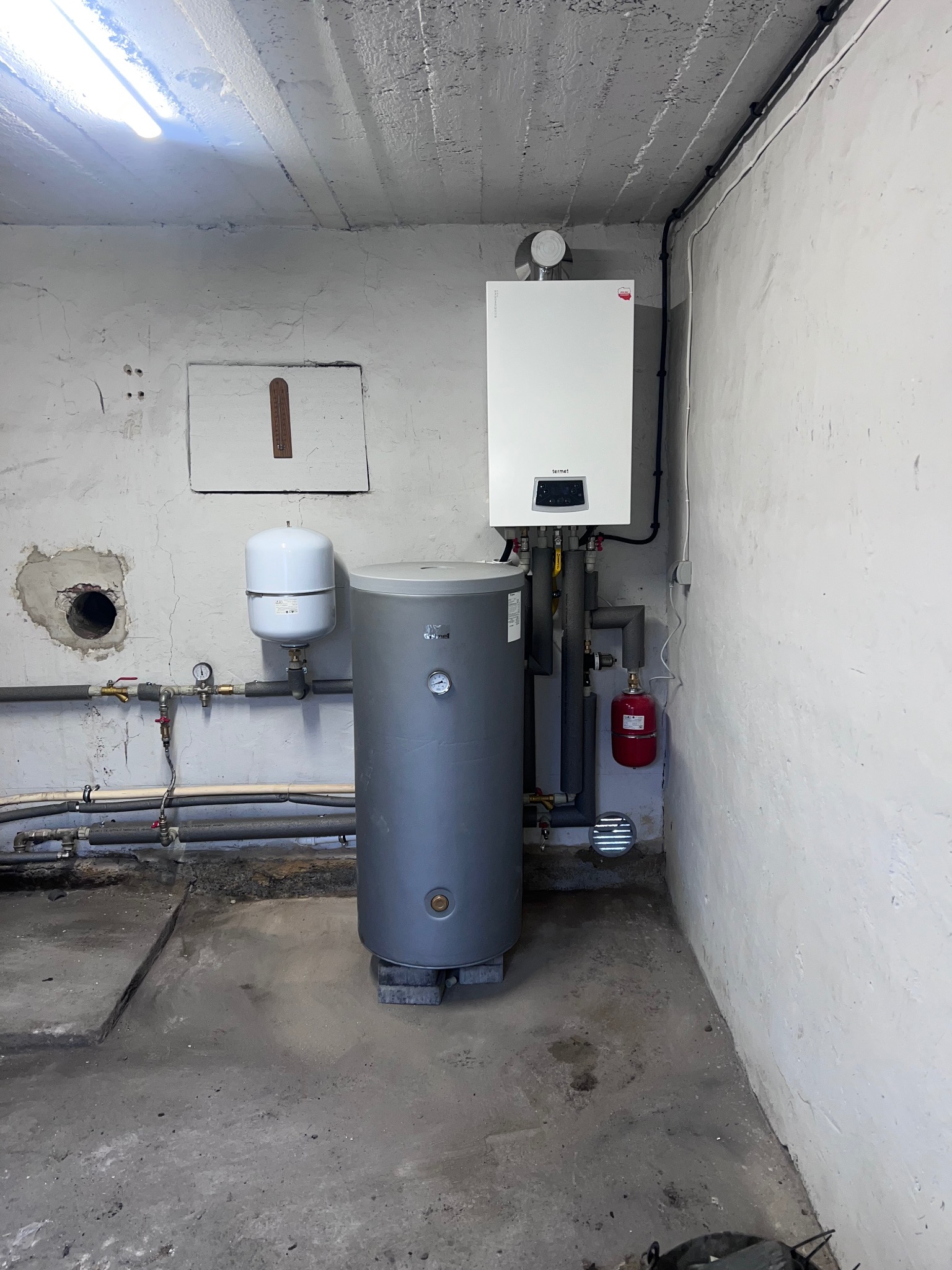 Biały piec gazowy Termet z szarym zasobnikiem wody i czerwoną butlą wyrównawczą w piwnicy z betonowym sufitem i instalacją hydrauliczną.