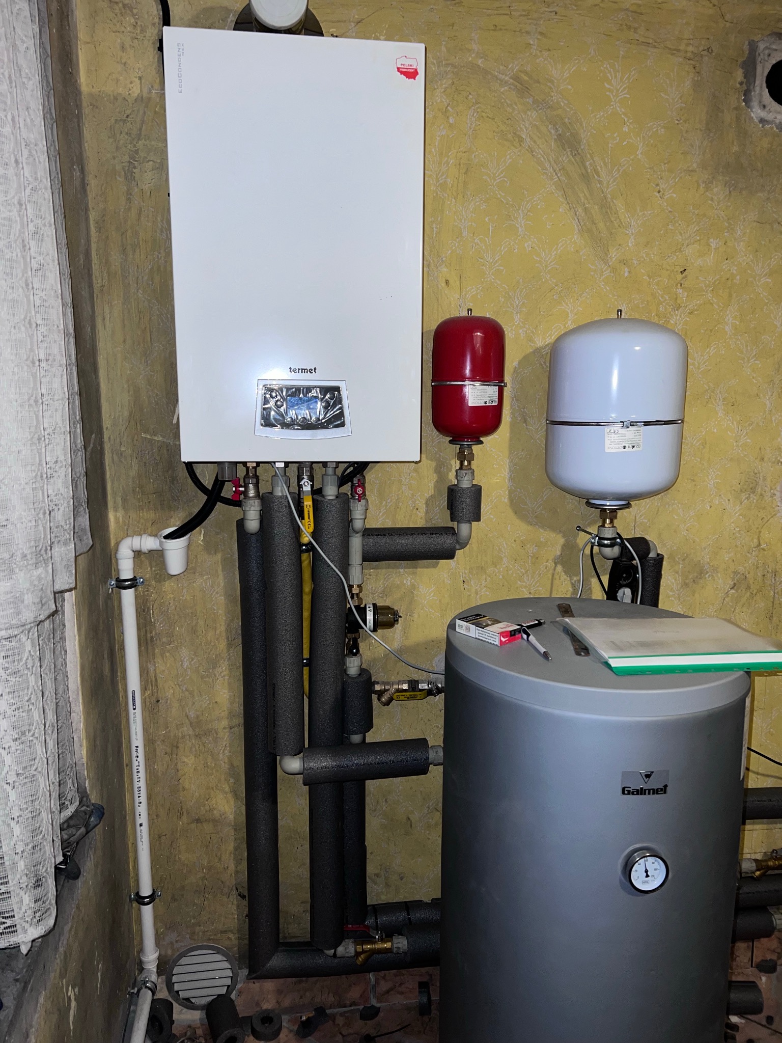 Biały piec gazowy Termet z szarym zasobnikiem Galmet, rury w izolacji, czerwony i biały zbiornik wyrównawczy na tle ściany w kwiaty. Widoczne narzędzia i dokumenty.