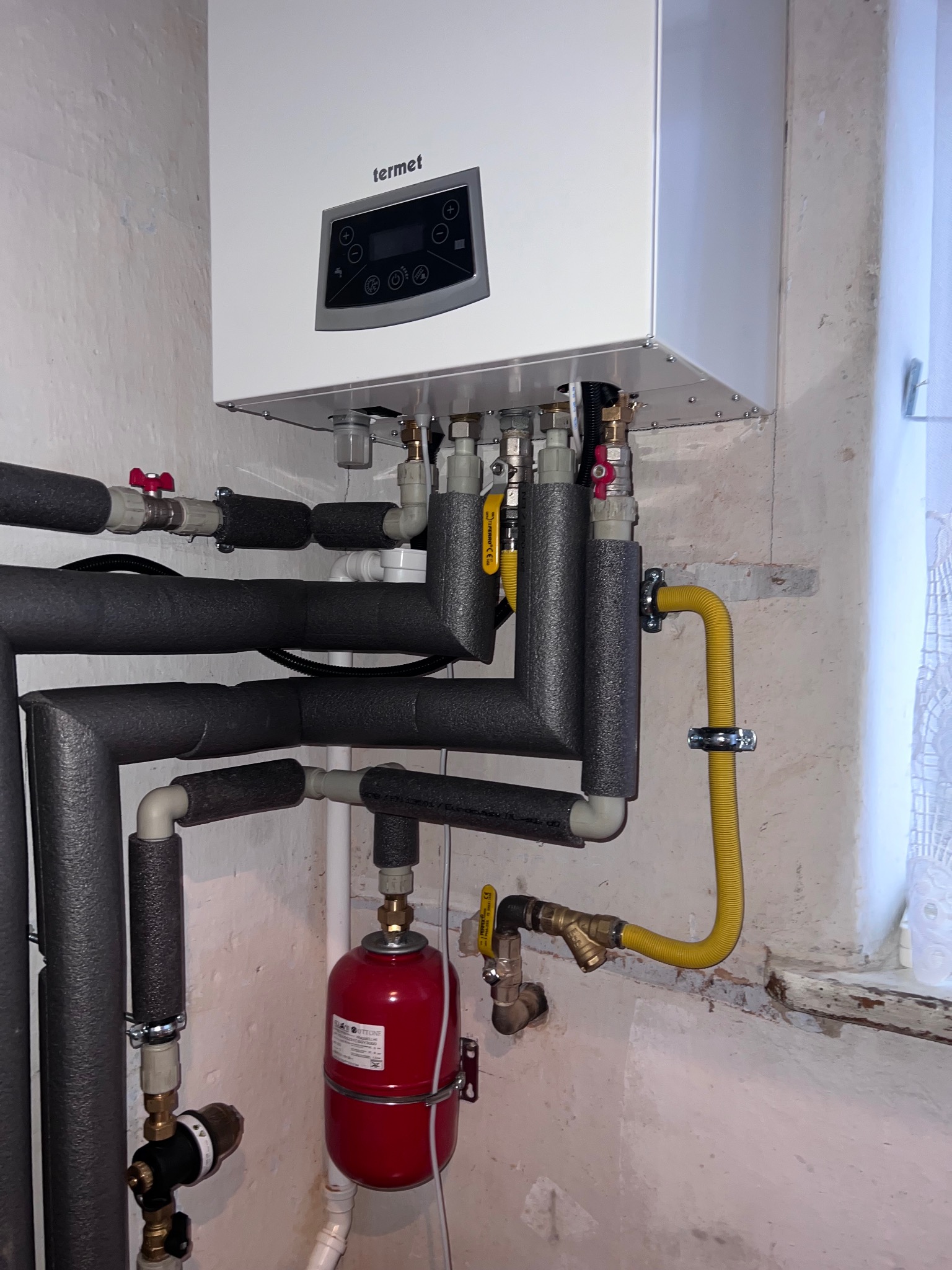 Biały piec gazowy marki Termet zamontowany na ścianie, z widocznymi rurami, zaworami i czerwonym zbiornikiem wyrównawczym. Instalacja grzewcza w piwnicy.