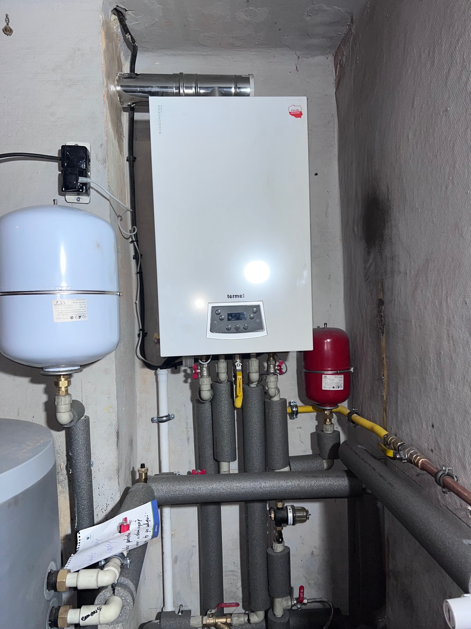 Biały piec gazowy Termet na ścianie z szarą instalacją, zbiornikiem i rurami w izolacji. Widoczne przyłącza, zawory i notatka przy rurach. Ściany surowe, z widocznymi śladami użytkowania.