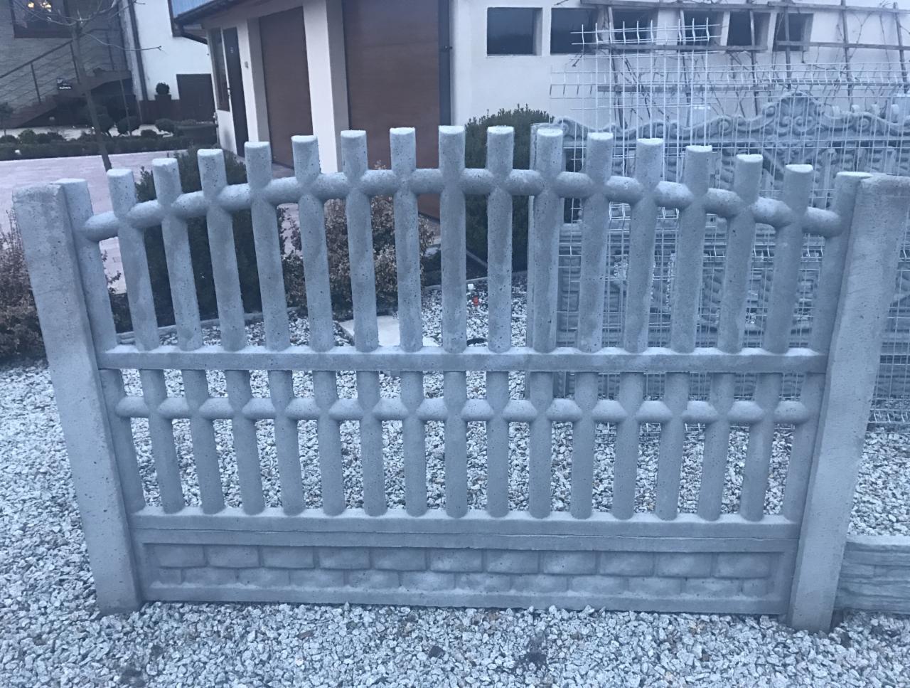 Szary, prefabrykowany element ogrodzenia z betonu o dekoracyjnym wzorze, leżący na żwirowym podłożu, z widocznymi śladami po produkcji. W tle dom z jasną elewacją i częściowo widoczne metalowe siatki.