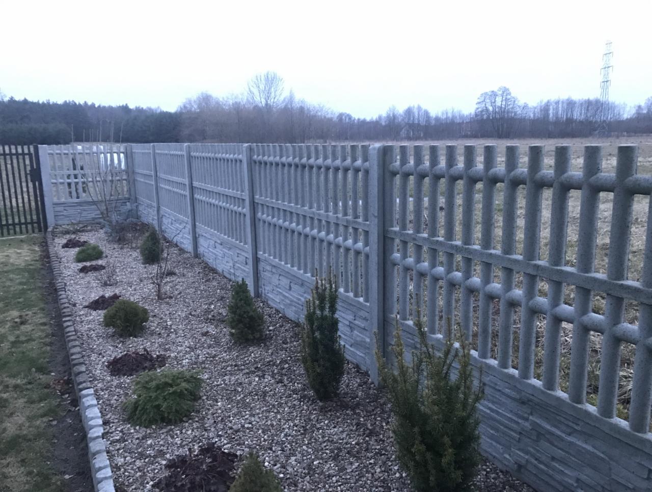Nowoczesne, szare ogrodzenie z prefabrykatów betonowych z poziomym wzorem imitującym kamień, oddzielające ogród od pola, z młodymi iglakami posadzonymi wzdłuż niego.