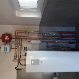 Kompletna instalacja grzewcza z białym kotłem, czerwonym i białym zbiornikiem wyrównawczym oraz miedzianymi rurami na ścianie z szarymi płytkami, widok z boku.