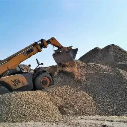 Żółta ładowarka teleskopowa JCB nabiera materiał sypki z dużej pryzmy pod błękitnym niebem.