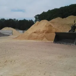 Żółta ładowarka kołowa stoi przed pryzmą jasnego kruszywa na placu składowym, w tle betonowe boksy z różnymi materiałami i zalesione wzgórze.