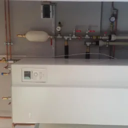 Nowoczesny, biały piec centralnego ogrzewania zamontowany na ścianie z widocznymi miedzianymi rurami instalacji i elementami armatury.