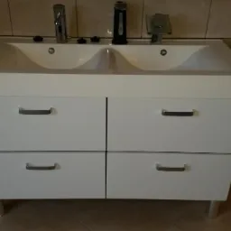 Biała szafka podwójna umywalkowa z czterema szufladami i chromowanymi uchwytami, widoczna na tle beżowych płytek.