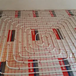 Białe rury ogrzewania podłogowego ułożone na folii z siatką w kolorze czerwonym, widoczne mocowania rur do podłoża.