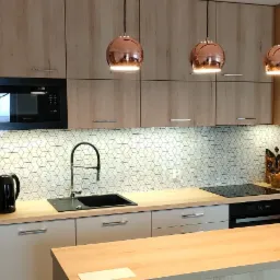 Nowoczesna kuchnia z drewnianymi frontami szafek, białymi płytkami w geometryczny wzór i miedzianymi lampami wiszącymi.