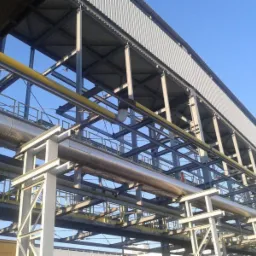 Zewnętrzny widok na stalową konstrukcję wsporczą z rurociągami, blacha trapezowa jako zadaszenie, błękitne niebo w tle.