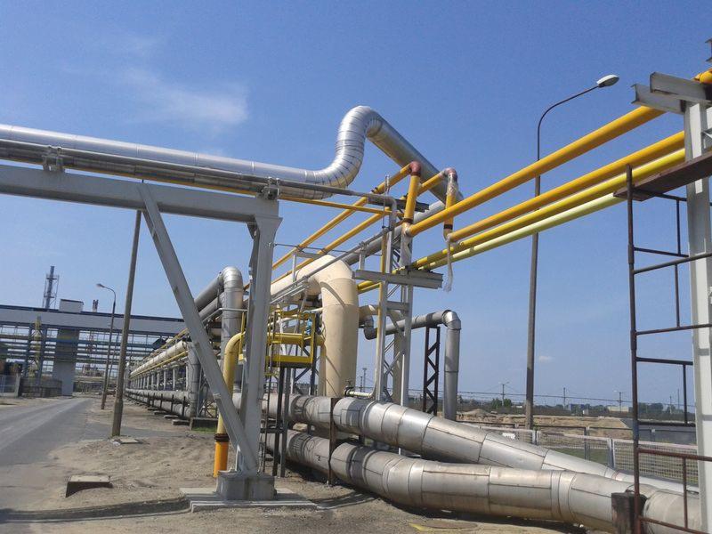 System rurociągów przemysłowych, rury w izolacji, wsparte na stalowej konstrukcji, z żółtymi rurami na górze, na tle błękitnego nieba i przemysłowego krajobrazu.