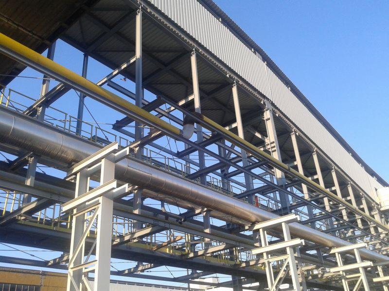 Zewnętrzny widok na stalową konstrukcję wsporczą z rurociągami, blacha trapezowa jako zadaszenie, błękitne niebo w tle.