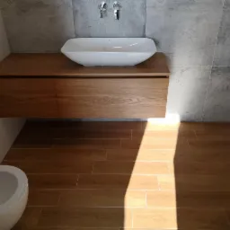 Nowoczesna łazienka z drewnianą szafką pod umywalką, białą umywalką nablatową i szarą ścianą z płytek imitujących beton, uzupełniona podłogą z płytek drewnopodobnych, oświetlona naturalnym światłem...