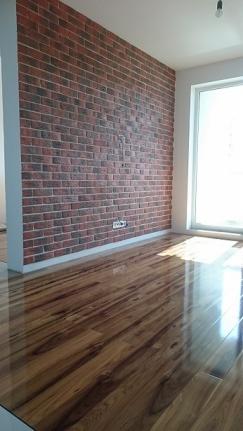 Wnętrze pokoju po remoncie: ściana wykończona tapetą imitującą czerwoną cegłę, błyszcząca podłoga z paneli drewnopodobnych, białe listwy przypodłogowe i ościeżnica okienna.