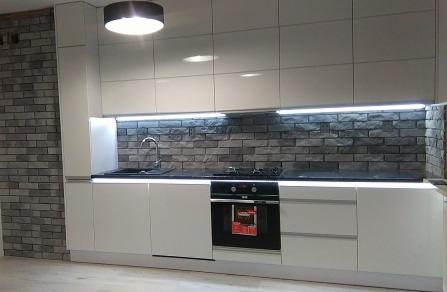 Nowoczesna kuchnia z białymi, lakierowanymi szafkami, czarnym blatem i ścianą wyłożoną szarą cegłą dekoracyjną, oświetlona listwami LED pod szafkami górnymi i dolnymi.