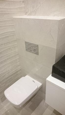 Nowoczesna toaleta podwieszana z białym siedziskiem i chromowanym przyciskiem spłukiwania na tle ściany wyłożonej płytkami imitującymi marmur z subtelną, falistą fakturą, obok umywalka z czarnym...