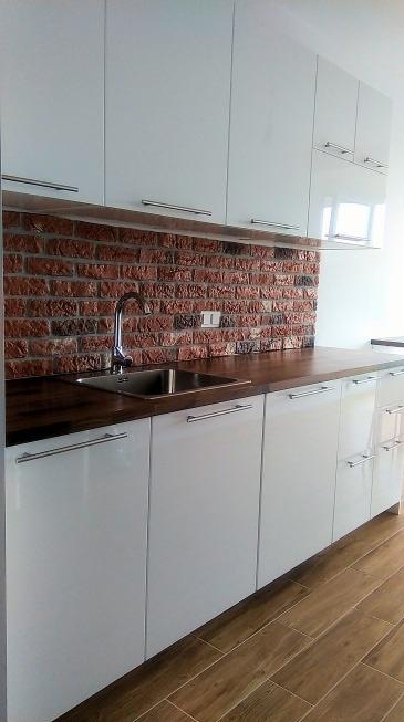 Kuchnia z białymi, błyszczącymi szafkami, drewnianym blatem i ścianą wyłożoną czerwoną cegłą. Widoczny stalowy zlew i bateria. Na podłodze panele imitujące drewno.
