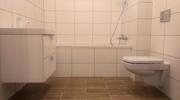 Wykończona łazienka z białymi płytkami ściennymi, brązowymi płytkami podłogowymi imitującymi drewno, białą zabudową wanny i umywalki oraz podwieszaną toaletą.