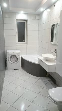 Jasna łazienka z narożną wanną, białą umywalką na szafce, pralką i kwadratowymi płytkami na ścianach i podłodze