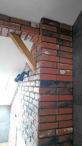 Wykończony fragment ściany z cegły klinkierowej, z widocznym drewnianym wzmocnieniem nad otworem, prawdopodobnie okiennym, z fragmentem skosu dachu w tle.