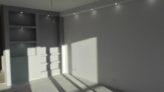 Pomalowane na szaro-biało wnętrze z oświetleniem punktowym w suficie i wnękami w ścianie, z widocznymi promieniami słonecznymi padającymi na podłogę.