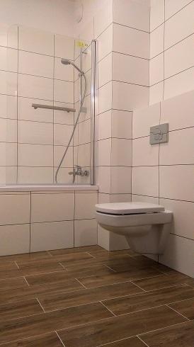 Nowoczesna łazienka z białymi płytkami ściennymi, drewnopodobnymi płytkami podłogowymi i podwieszaną toaletą.